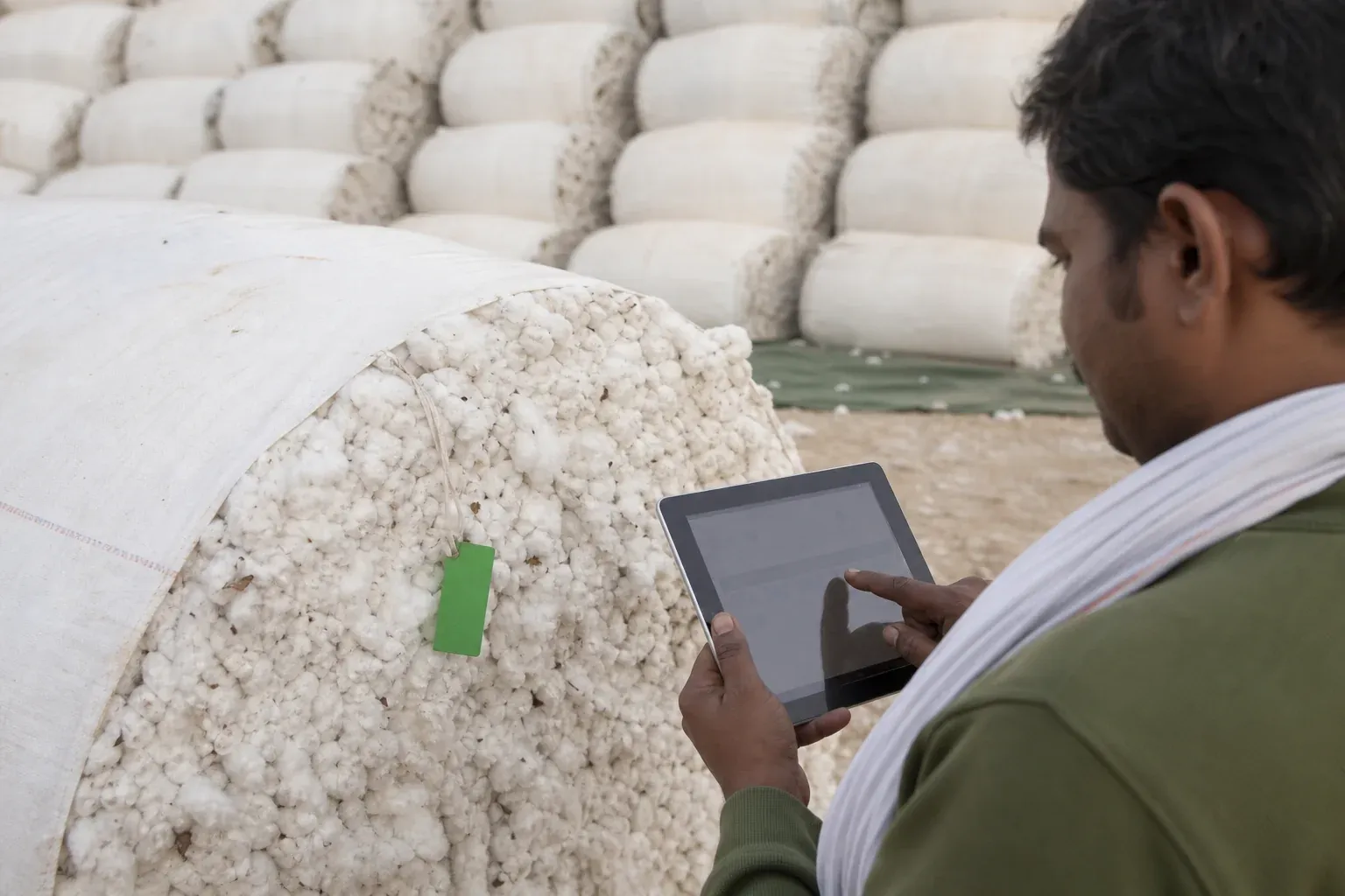 Photorealistic photo of a field technician using a tablet to scan and log a tagged cotton bale at a rural ginning yard in India, hands visible on the tablet screen showing a simple tracking interface, cotton bales stacked in background