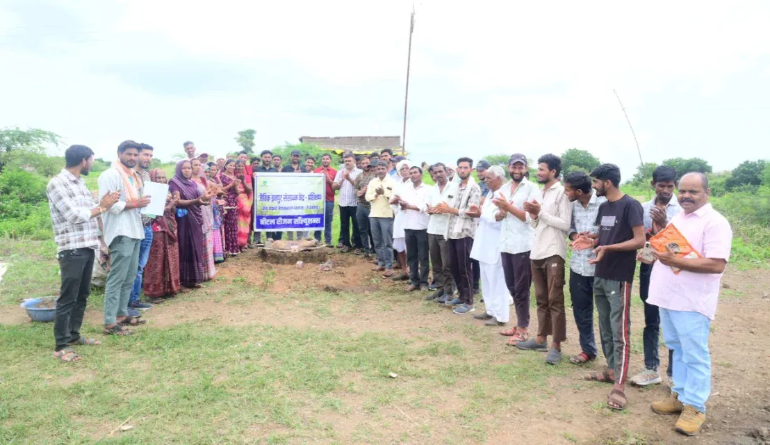 Outdoor event photo of a large group of villagers, men and women, standing in a semicircle around a small cleared patch of earth in an open rural field and applauding. A banner at the center is labele